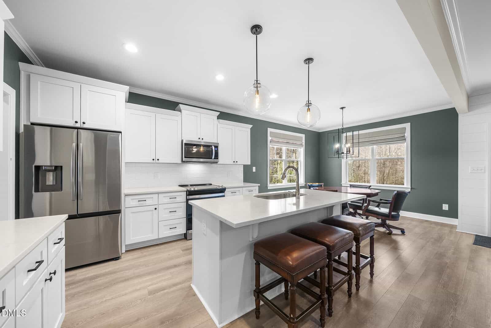 Modern kitchen with white cabinets, stainless steel appliances, green accent walls, large island with sink, and three brown leather barstools. Dining area with table and chairs near large windows overlooking trees.