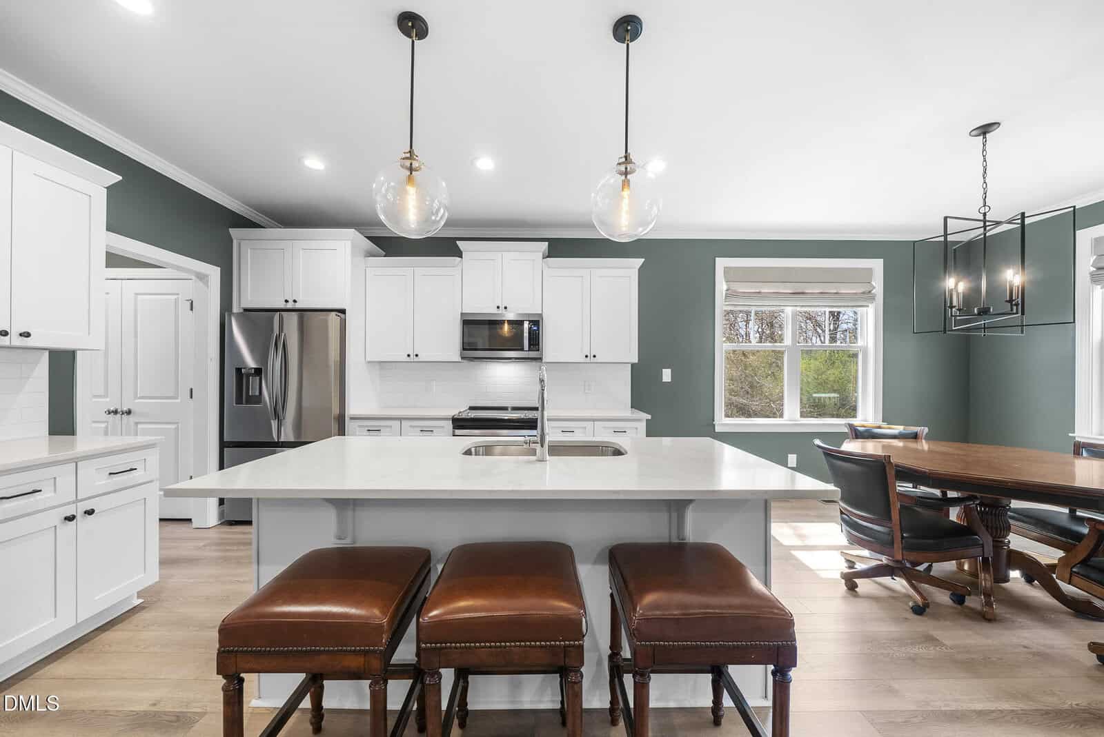 Modern kitchen with white cabinets, stainless steel appliances, a large island with sink, three brown leather stools, green walls, and adjacent dining area with window and chandelier.