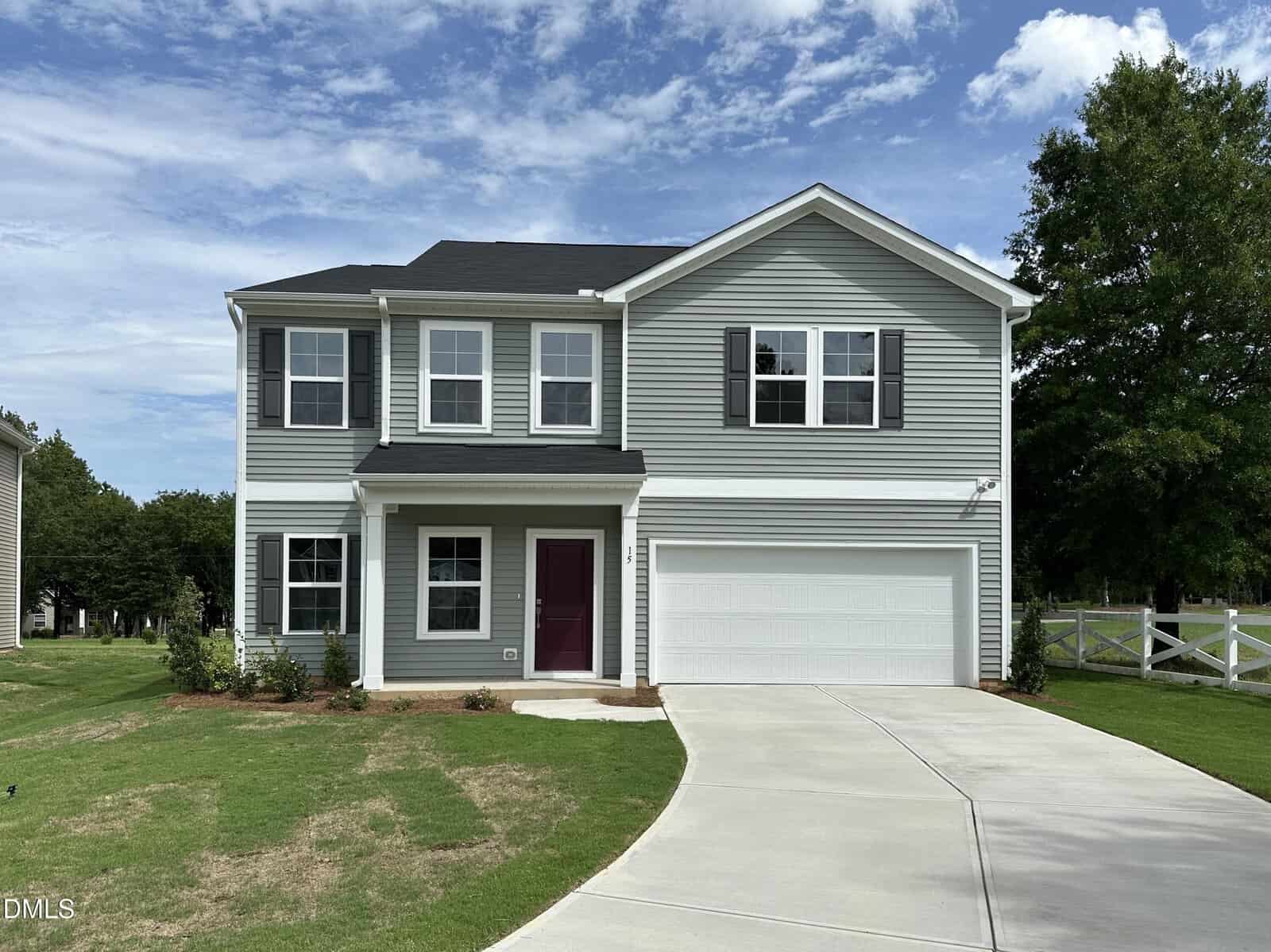 Two-story gray house with white trim, black roof, and attached two-car garage located at 15 Buckthorn Drive, Youngsville. A concrete driveway curves to the garage. The front yard features green grass and small plants near the entrance, with trees and a white fence in the background.