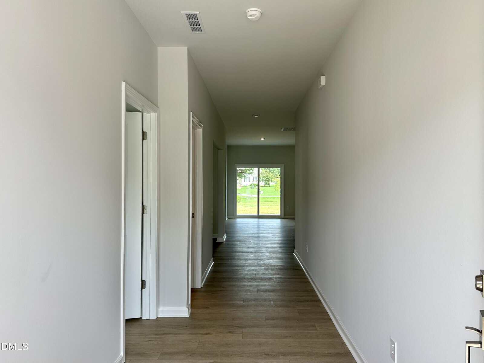 A long hallway with light-colored walls and wood flooring, leading to a glass door that opens to a grassy backyard. Two open doorways are on the left side of the hallway.