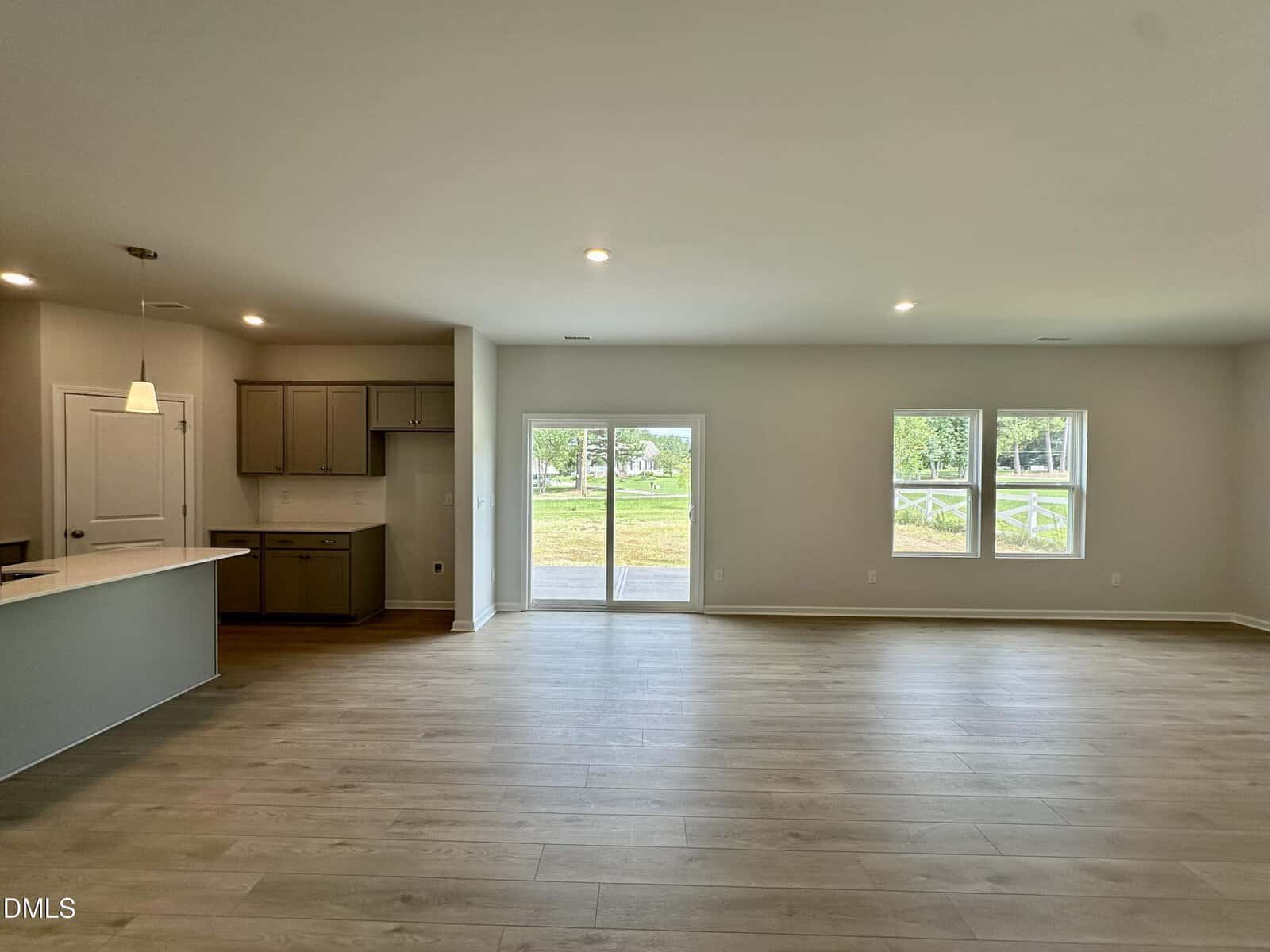 Spacious, empty living area with light wood floors, large windows, and sliding glass doors. Kitchen with beige cabinets and pendant light is visible to the left. Natural light fills the room.