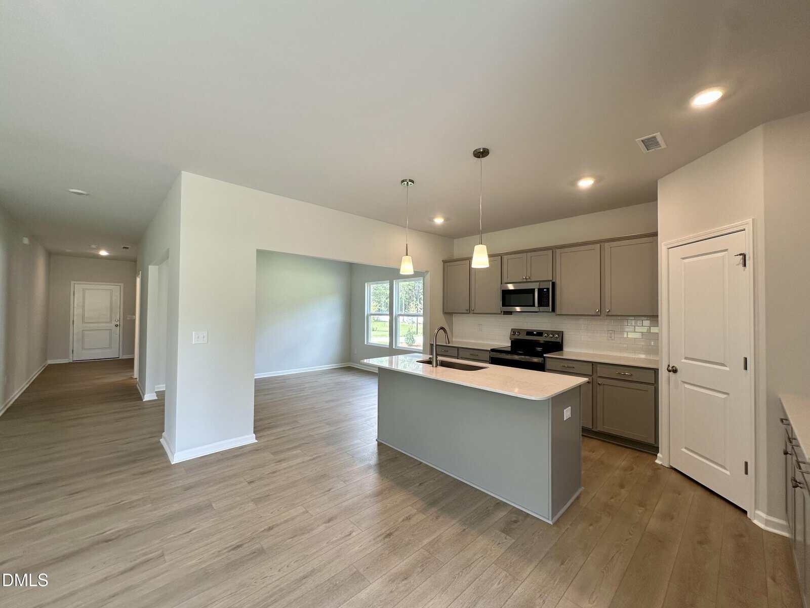 Modern kitchen with light wood floors, gray cabinets, stainless steel appliances, pendant lights above a center island, and a bright open layout with large windows in the adjacent dining area.