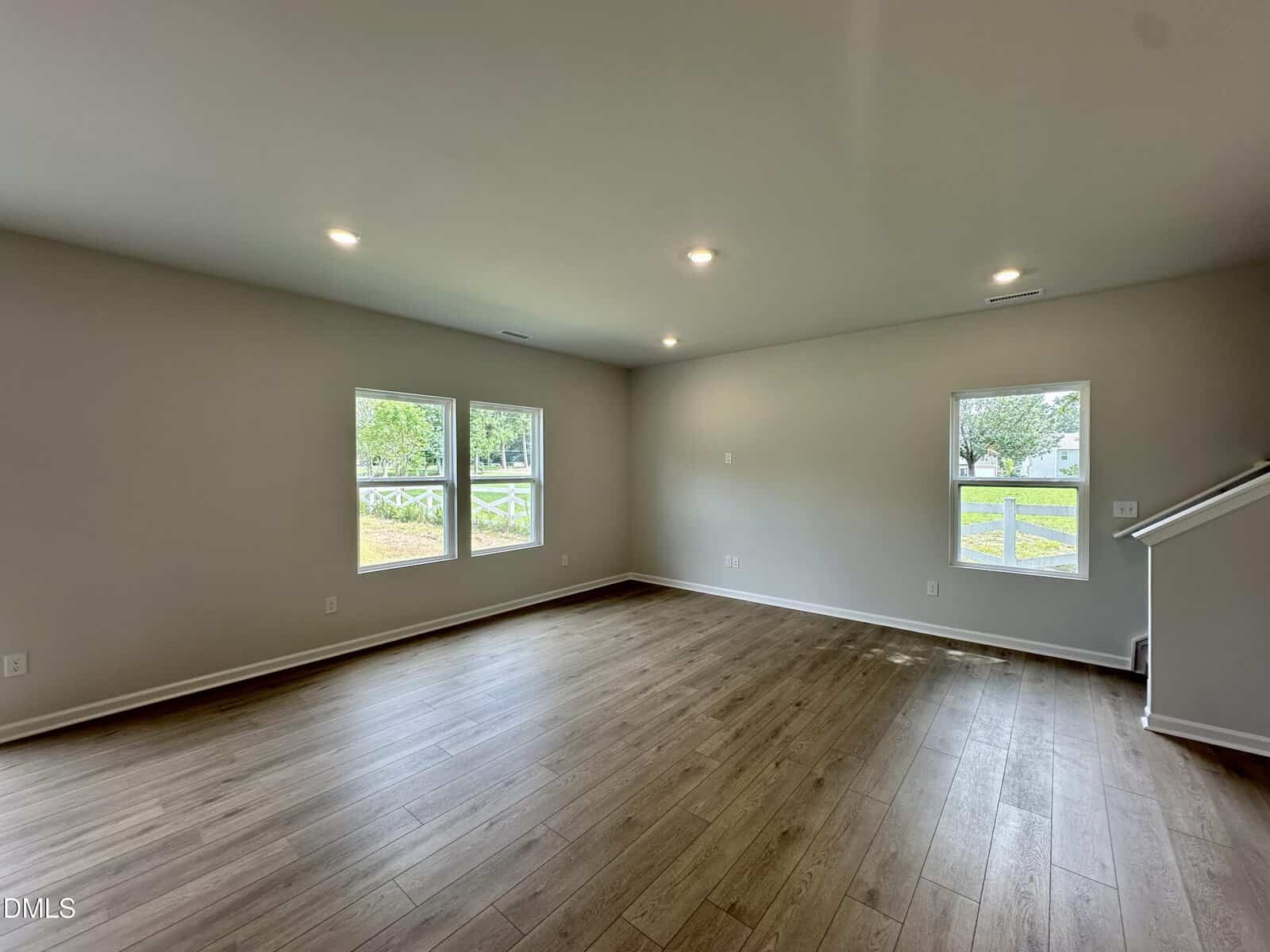 Spacious, empty living room with light wood floors, beige walls, recessed lighting, and multiple windows letting in natural light.