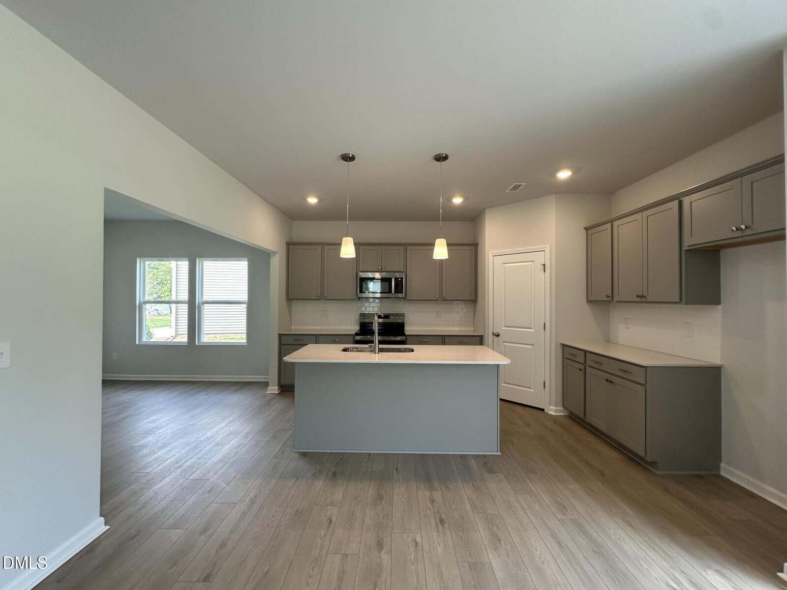 Modern kitchen with gray cabinets, a center island, pendant lights, stainless steel appliances, and light wood flooring. The kitchen opens to a dining area with large windows. Walls are painted light beige.