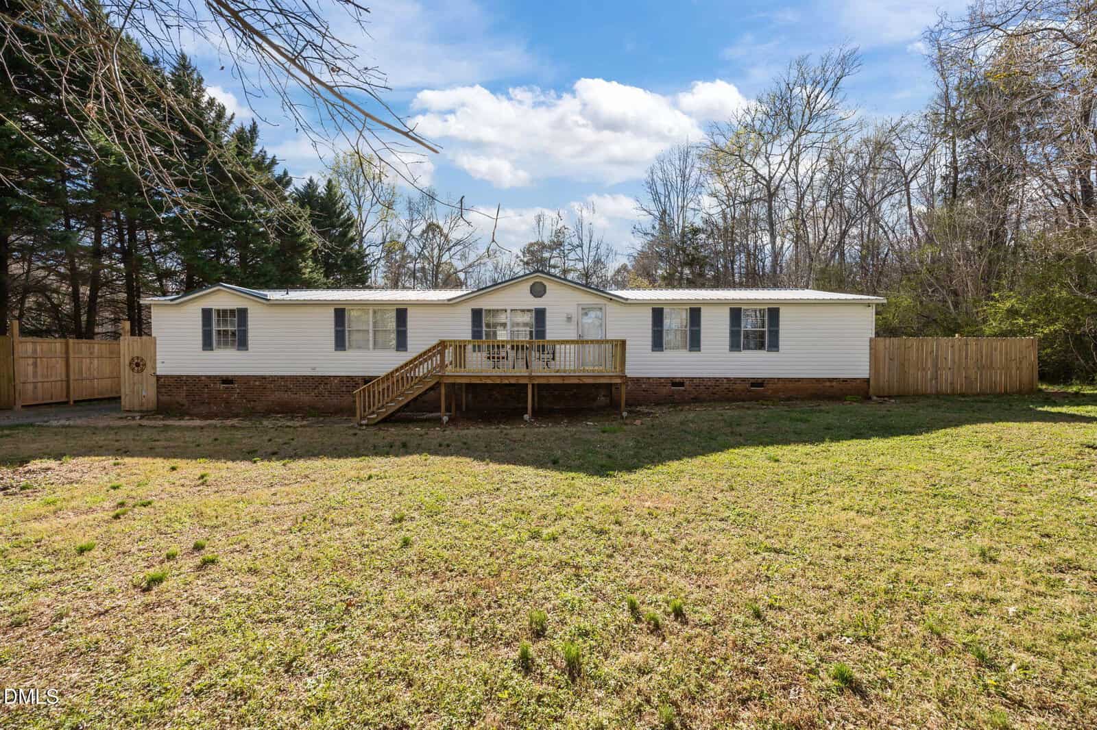 A single-story manufactured home with white siding, a brick foundation, and a wooden front porch is set on a grassy lawn. There is a wooden fence on both sides and trees in the background under a partly cloudy sky.