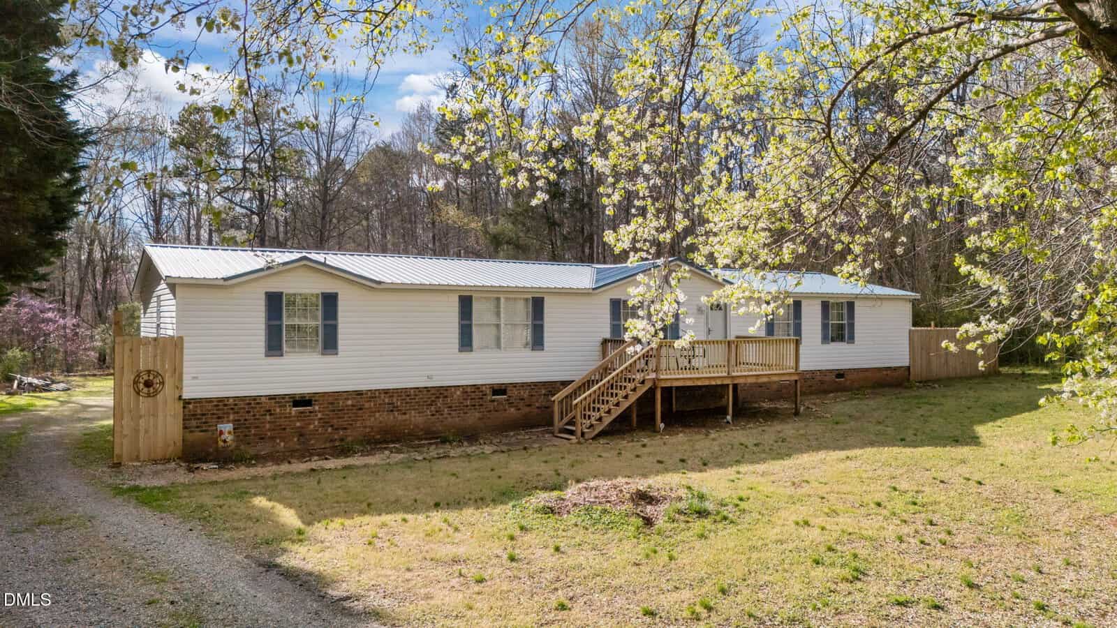 A single-story white manufactured home with a brick foundation sits in a grassy yard with trees in the background. Wooden steps lead to a small front porch, and a gravel driveway runs along the side.