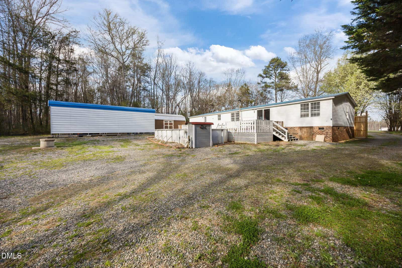 A white manufactured home on a large, gravel-covered lot with a small above-ground pool, trees in the background, and a blue-and-white shed to the left under a partly cloudy sky.
