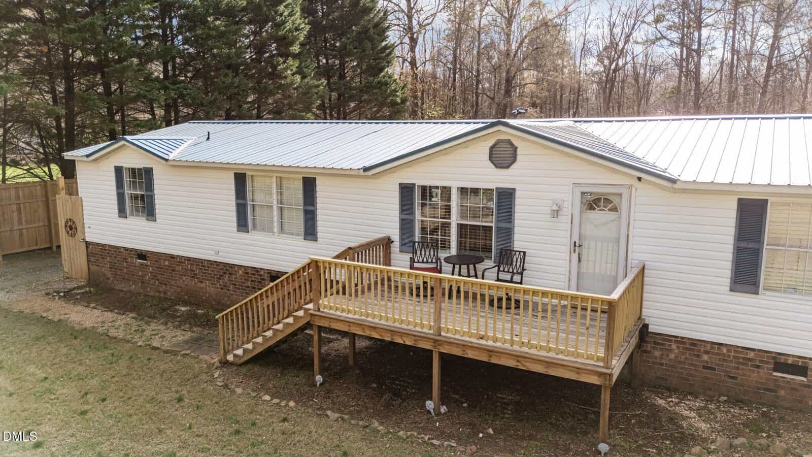A white manufactured home with a brick foundation, a metal roof, and a wooden front porch with two chairs and a small table, surrounded by trees and grass.