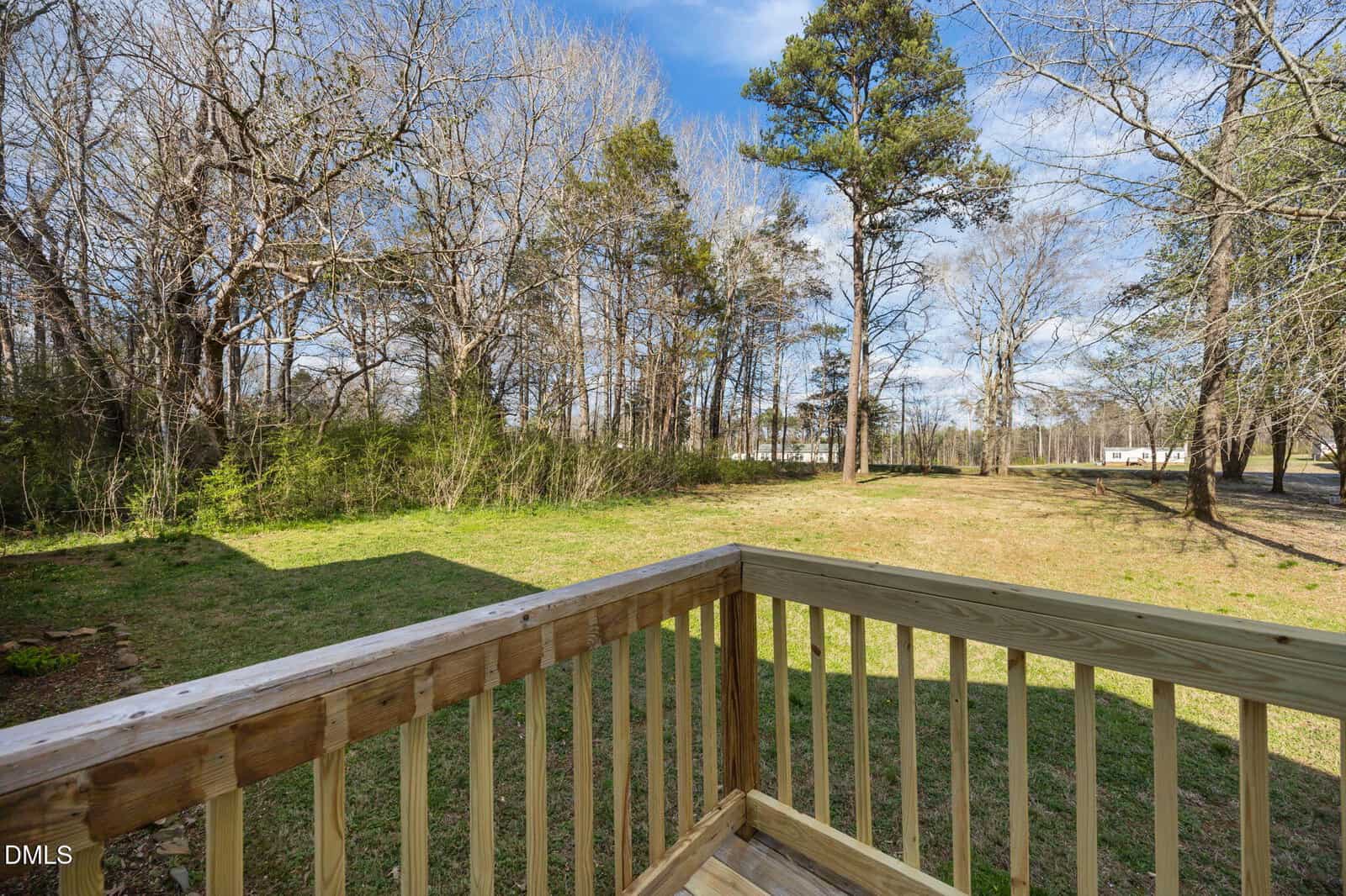 View from a wooden deck looking out onto a grassy yard bordered by tall trees under a partly cloudy blue sky.