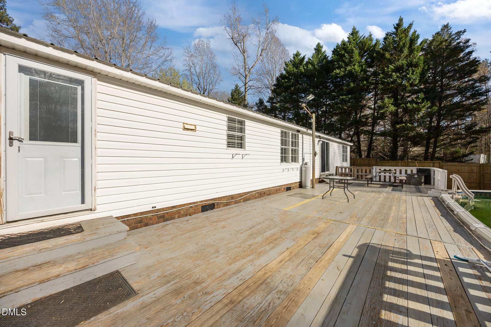 A spacious wooden deck attached to a white mobile home, surrounded by trees under a blue sky with scattered clouds. Outdoor seating and a small table are visible on the deck.