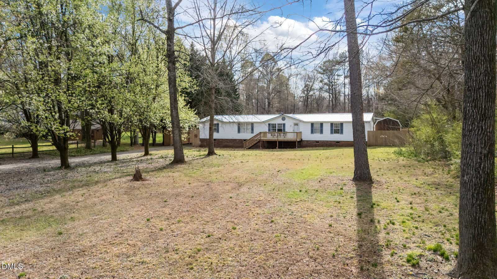 A white manufactured home with a raised wooden front porch sits on a large, grassy lot surrounded by trees under a partly cloudy sky.