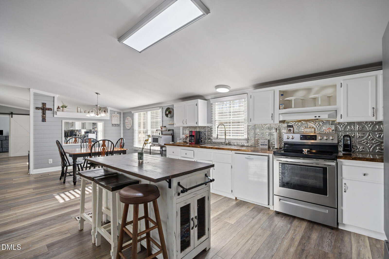 Modern farmhouse kitchen with white cabinets, stainless steel appliances, a wooden island with stools, light wood floors, and a dining area in the background. Sunlight enters through multiple windows, creating a bright, airy feel.