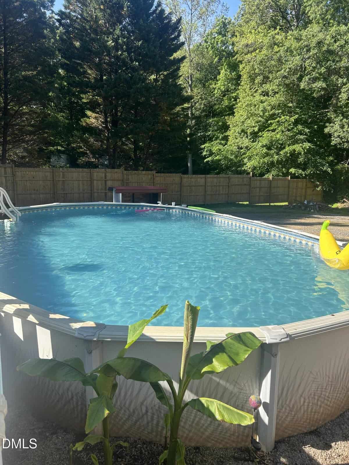 Enjoy this above-ground backyard pool at 9089 Grassy Creek Road, Bullock, surrounded by a wooden fence and trees. A banana plant adds a tropical touch, while a yellow inflatable duck floats nearby and a bench awaits at the far end.
