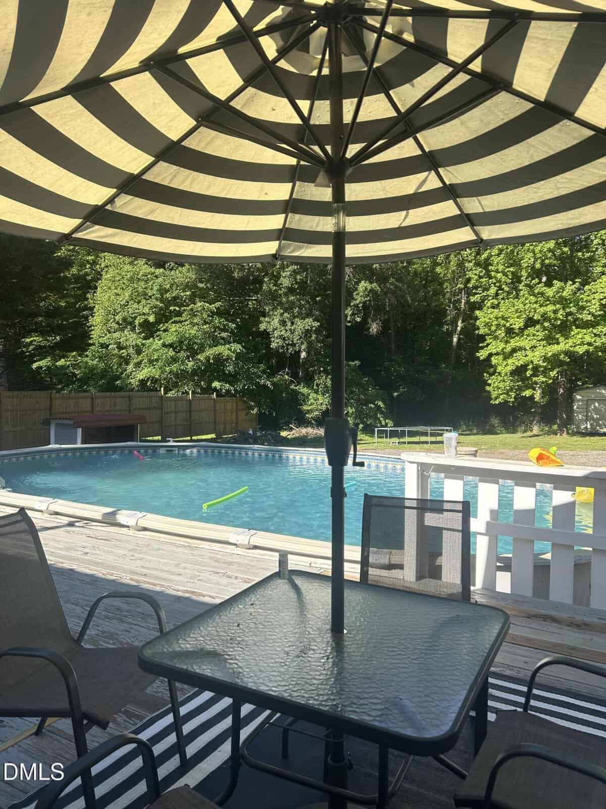 A shaded patio table with chairs sits under a large striped umbrella beside a clear outdoor swimming pool, surrounded by a deck and trees in the background. Pool toys float in the water.