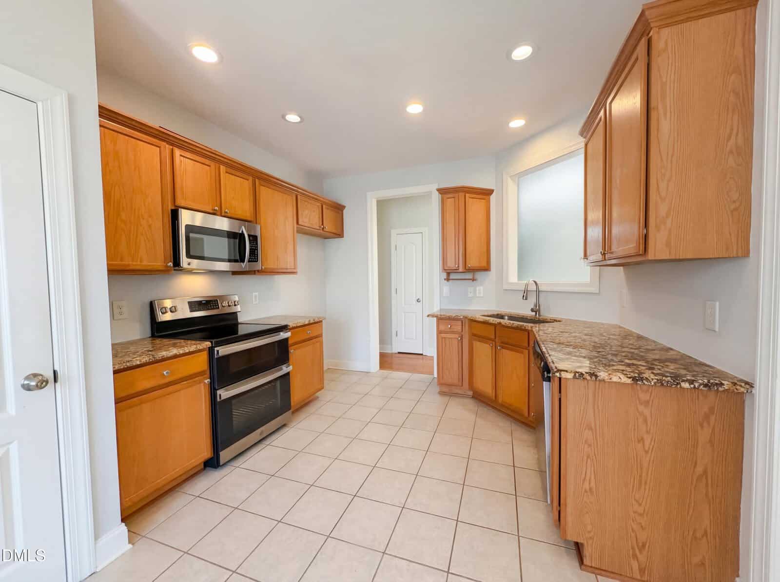 A kitchen with light brown wooden cabinets, stainless steel appliances, brown granite countertops, tiled floor, recessed ceiling lights, and a large window above the sink.