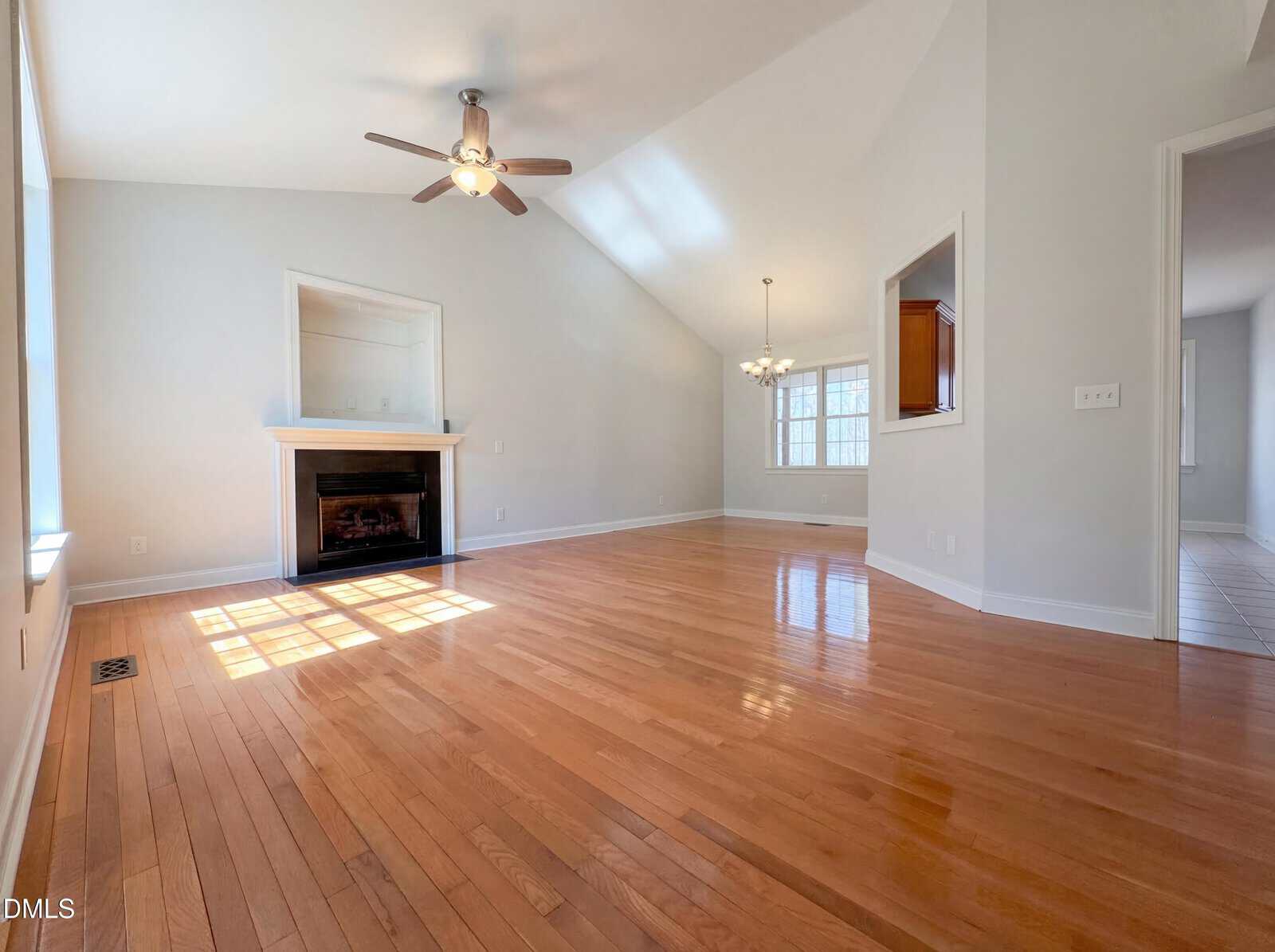 Spacious empty living room with hardwood floors, vaulted ceiling, ceiling fan, fireplace, and large windows letting in natural light. An open doorway leads to an adjacent room with tile flooring.