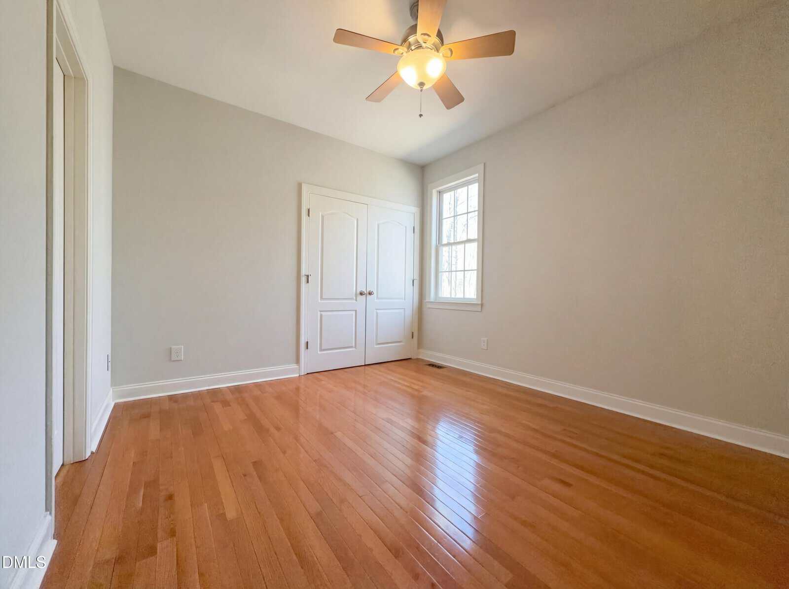 An empty room with light-colored walls, a ceiling fan with a light, a window, double white doors, and a wooden floor. Natural light enters through the window, and the room appears clean and well-maintained.