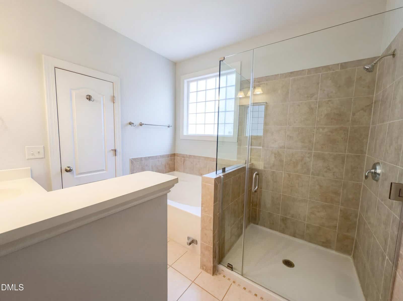 A bathroom with a glass-enclosed shower, beige tile walls, a built-in bathtub, a window above the tub, a towel rack, and a white door. The walls and floor are light-colored.