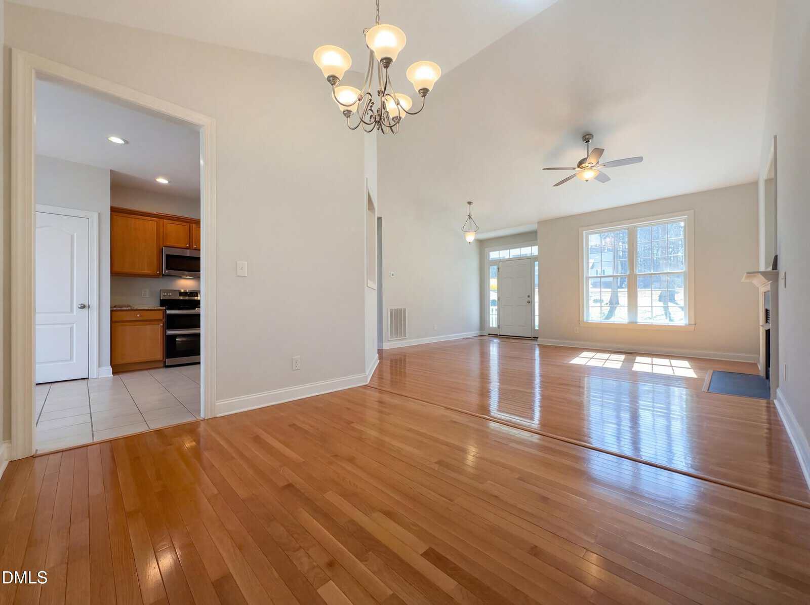 A spacious open-plan living area with light wood floors, a chandelier, ceiling fan, and large windows. The kitchen with wooden cabinets and stainless appliances is visible through a doorway on the left.