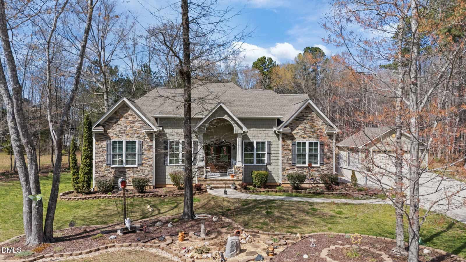 A single-story house with a stone and siding exterior, arched entrance, and gable roof, surrounded by leafless trees. A curved path and small garden are in front, and a detached garage is visible on the right.