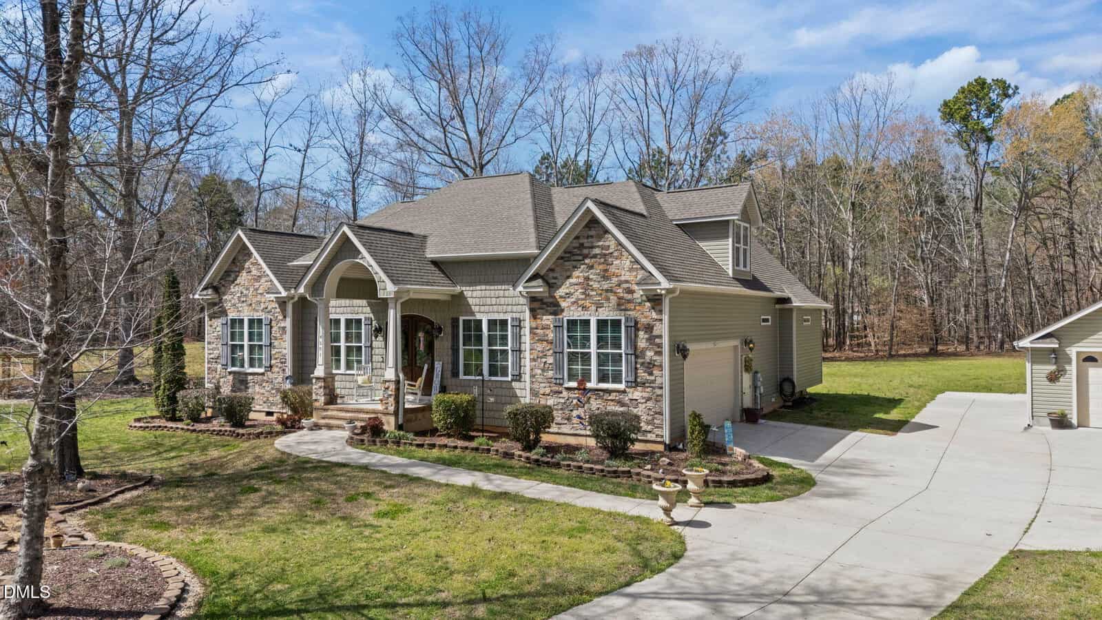 A large two-story house with a stone and siding exterior, multiple gables, white columns, and a curved walkway, surrounded by grass, shrubs, and trees. A detached garage sits to the right of the driveway.