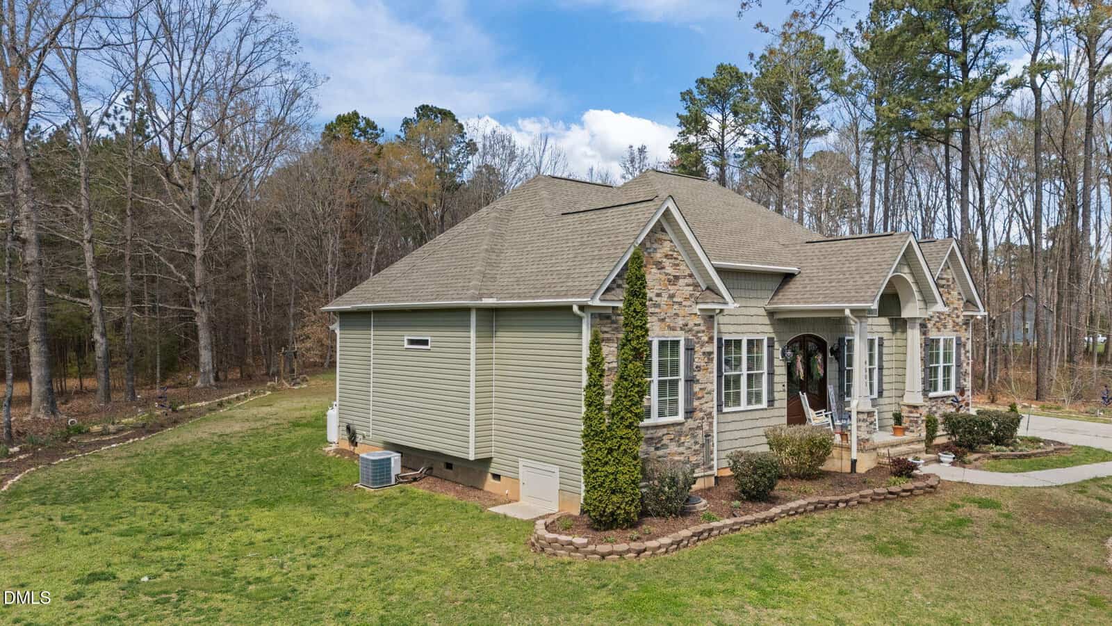 A single-story house with light green siding, stone accents, and a gabled roof sits on a grassy lawn surrounded by trees. There are shrubs and minimal landscaping near the front entrance.