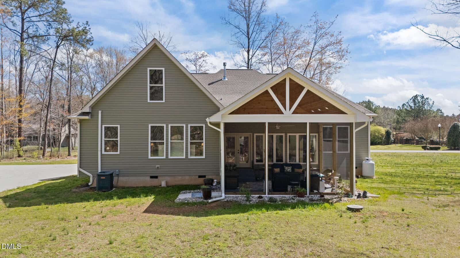 A gray house with a gabled roof features screened-in back porch, outdoor furniture, lawn, and surrounding trees under a partly cloudy sky. The house is located on a corner lot with a road visible on the left.