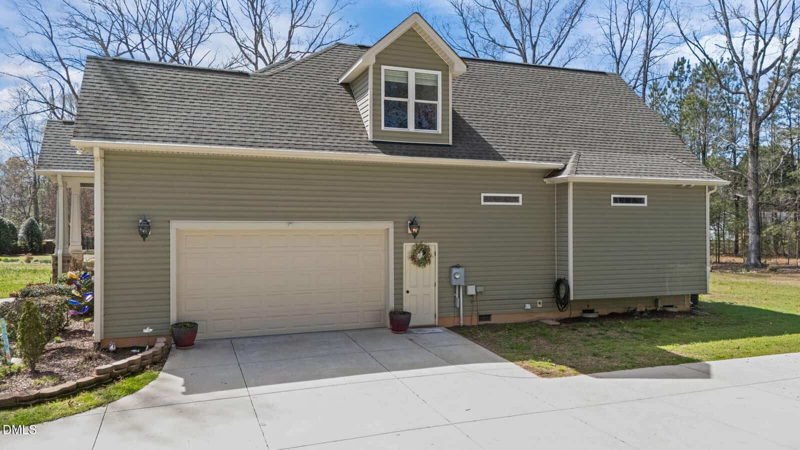 A two-story house with green siding, a two-car garage, small dormer window above, concrete driveway, potted plants near the entrance, and trees in the background.