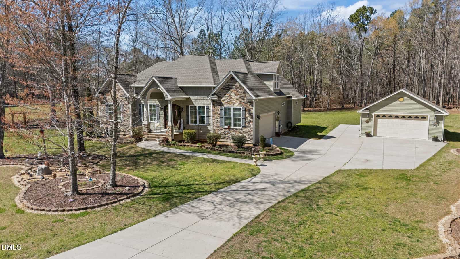 A two-story house with stone and siding exterior, landscaped front yard, curved concrete driveway, detached two-car garage, and surrounded by trees under a partly cloudy sky.