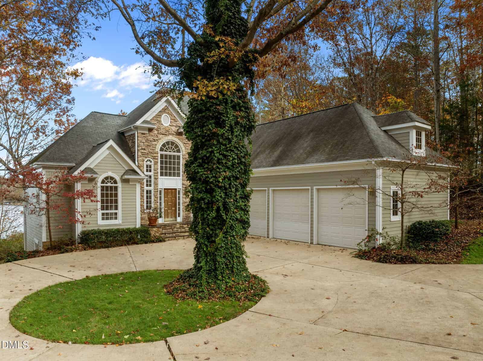A two-story house with light gray siding, stone accents, and three white garage doors sits behind a circular concrete driveway. A large tree with ivy stands in the center of the driveway, surrounded by autumn foliage.