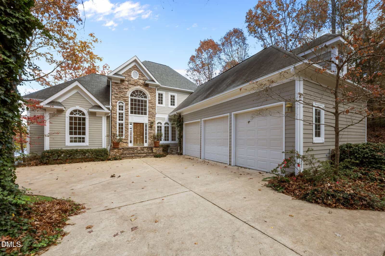 Two-story suburban house with stone and siding exterior, large arched windows, a three-car garage, and a wide driveway. Trees with autumn foliage surround the property.