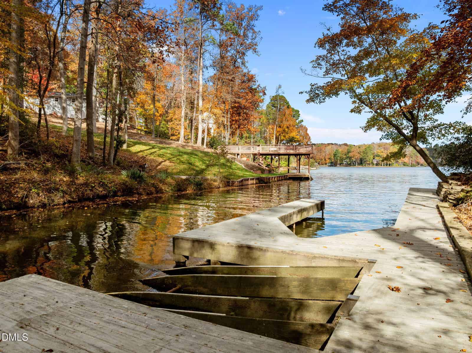 A wooden dock with boat slips extends into a calm lake surrounded by trees with autumn foliage; a wooden deck overlooks the water, and houses are partly visible among the trees in the background.