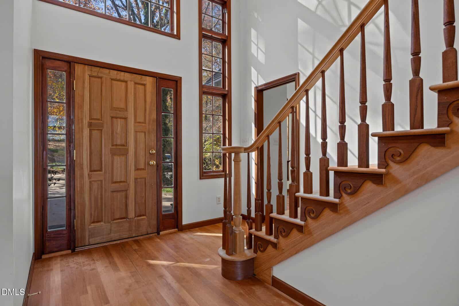 A wooden front door with side windows opens to a foyer with hardwood floors and a staircase featuring turned balusters, decorative trim, and natural light streaming through tall windows.