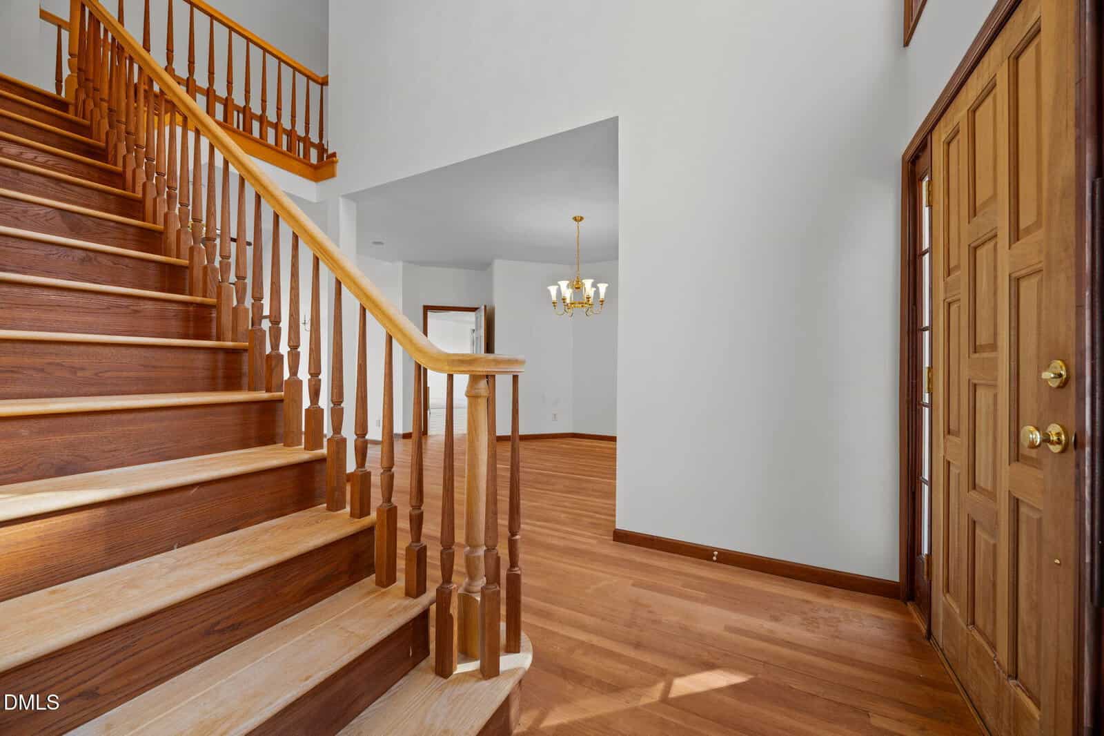 A wooden staircase with a matching handrail is next to a wooden front door. The entryway leads to an open room with wood floors, white walls, and a gold-tone chandelier hanging from the ceiling.