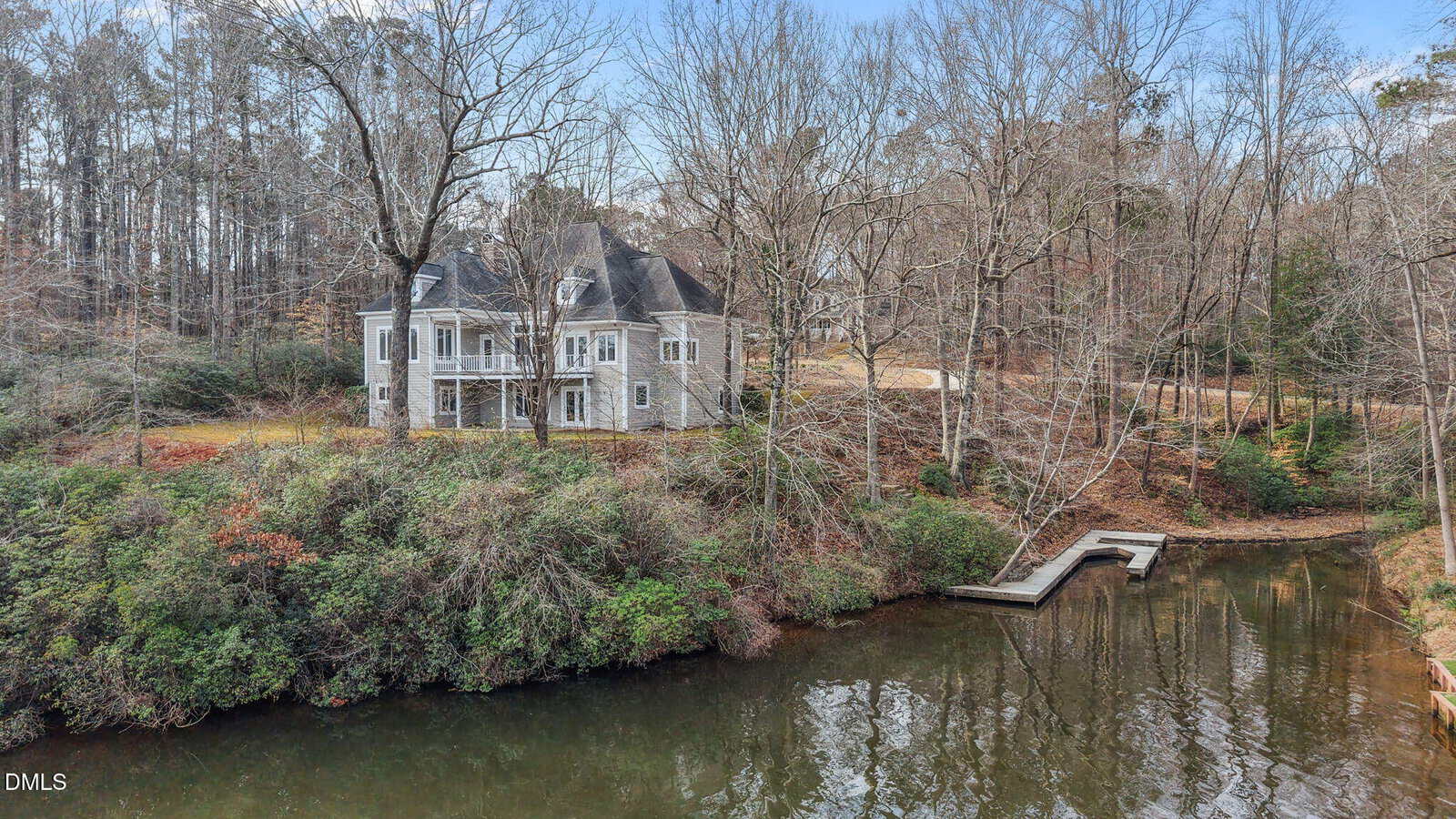A stone house with multiple gables sits on a wooded lot by a calm pond. A wooden dock extends into the water. The trees are leafless, suggesting late fall or winter.