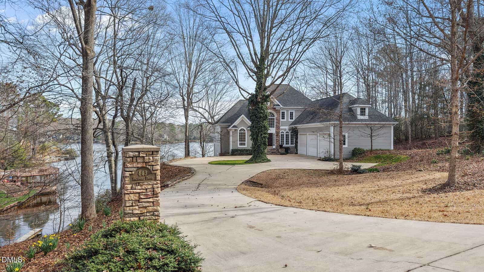 A two-story house with a three-car garage sits at the end of a curved driveway beside a lake. Leafless trees surround the property, and a stone mailbox stands near the driveway entrance.