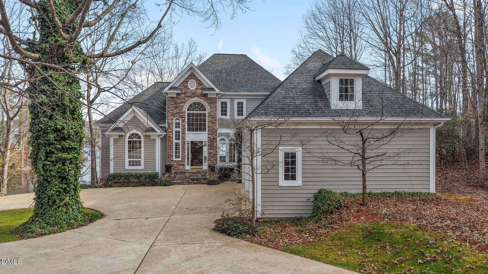 A two-story gray house with white trim, multiple gables, stone accents around the front entrance, large windows, and a circular driveway, surrounded by leafless trees on a wooded lot.