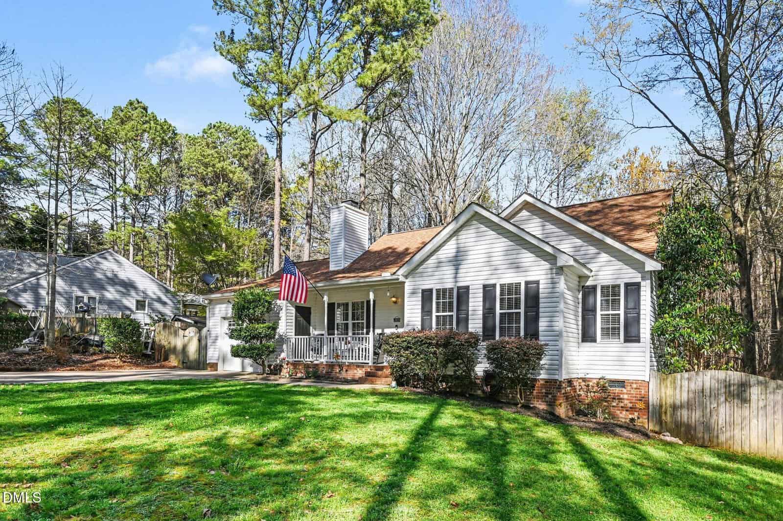 A single-story white house with brown roof, front porch with American flag, surrounded by green lawn, shrubs, trees, and neighboring homes under a partly cloudy sky.