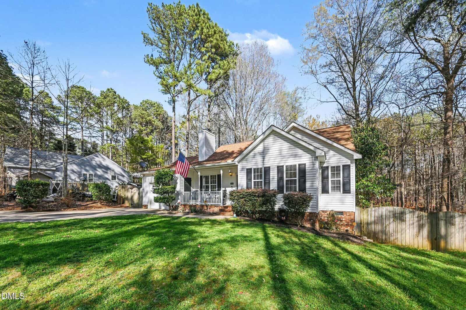 A single-story white house with a front porch and American flag, surrounded by trees and a large green lawn, sits in a suburban neighborhood on a sunny day.