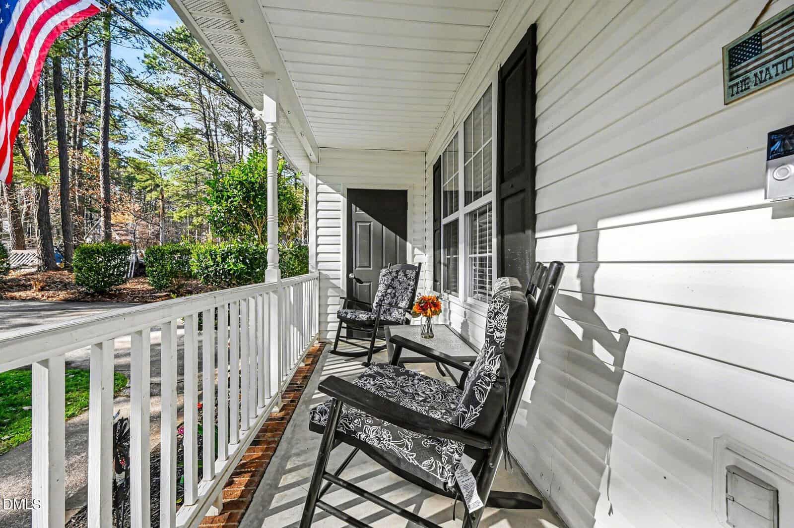 A white house porch with two cushioned chairs, a small table with a flower arrangement, black shutters, and an American flag. Trees and bushes are visible in the background.