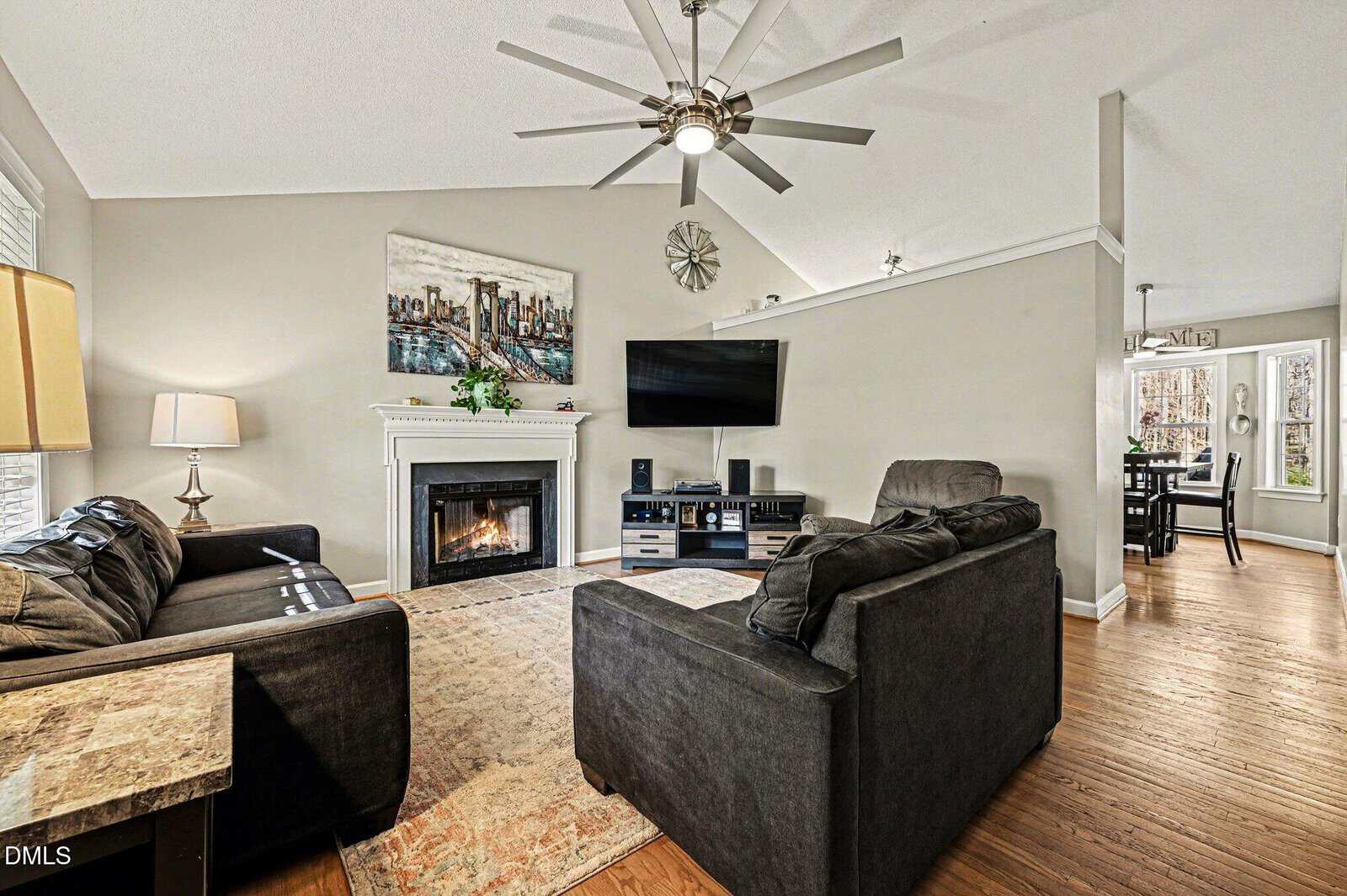 A living room with two dark sofas, a rug, a fireplace with a painting above it, a mounted TV, media console, large ceiling fan, wooden floors, and a partial view of a dining area in the background.