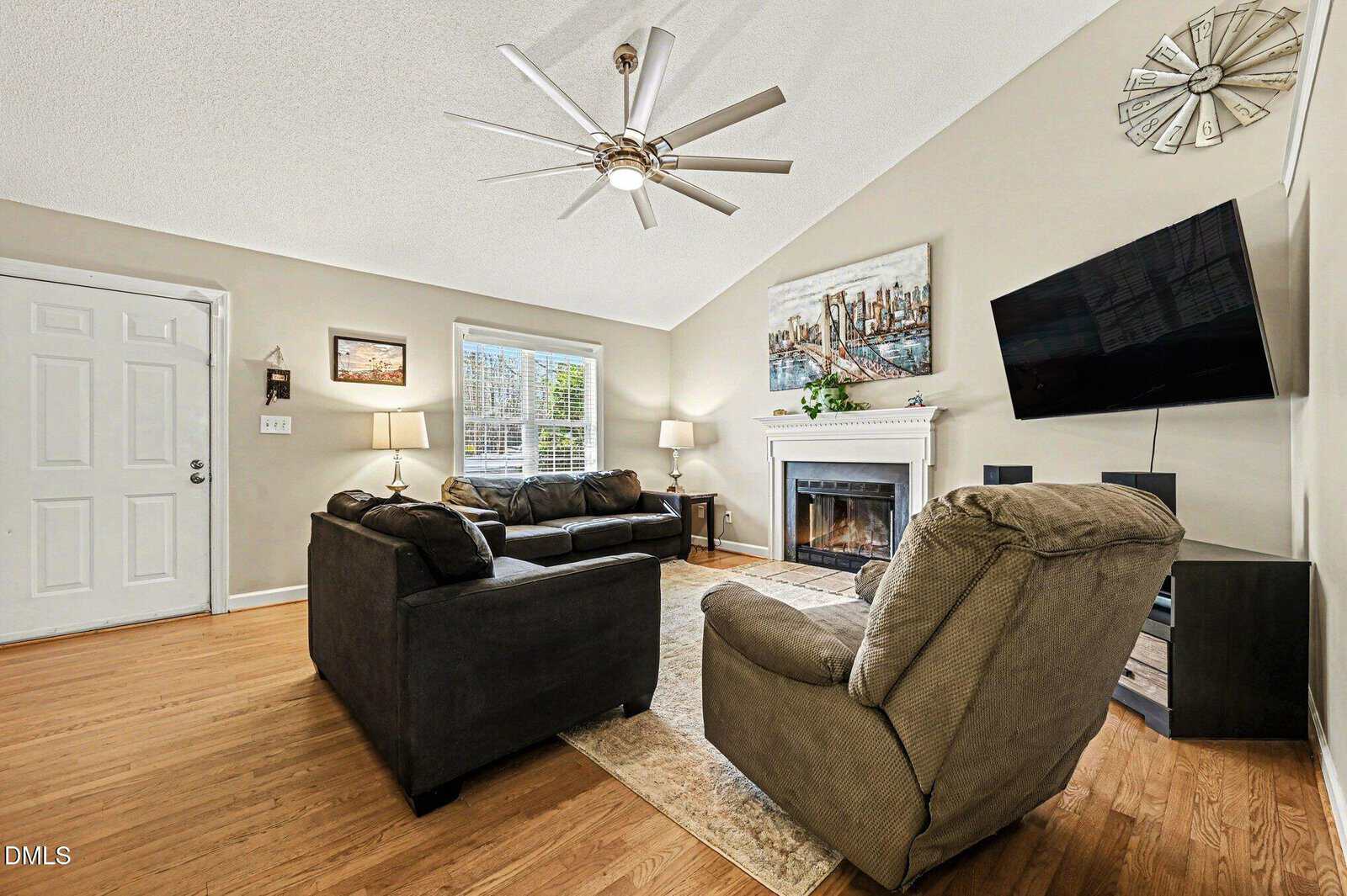 A living room with hardwood floors, a gray sofa set, an armchair, a fireplace, a wall-mounted TV, and a ceiling fan. Light walls, large windows, and decorative items are present. The front door is visible on the left.