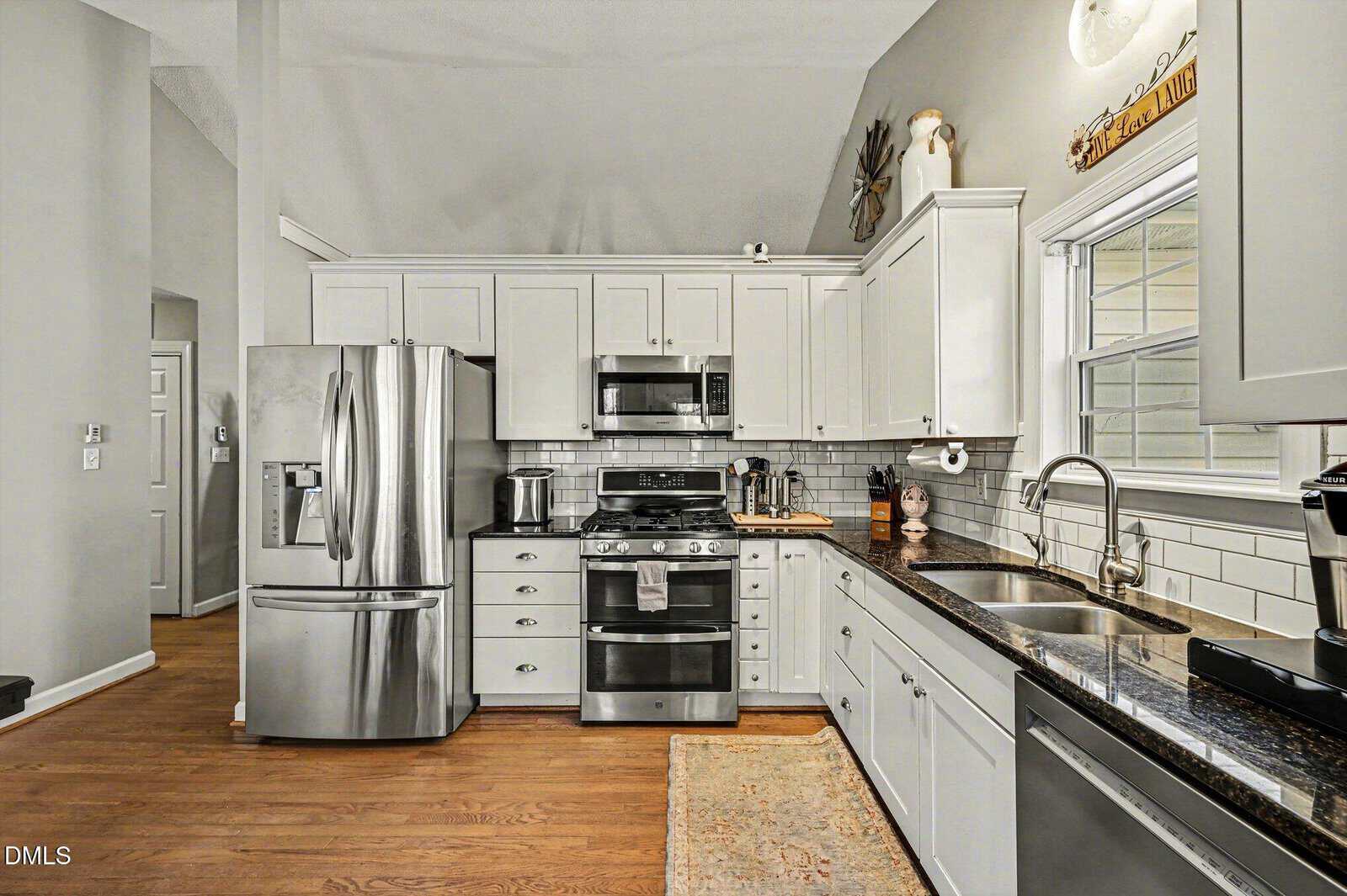 Modern kitchen with white cabinets, stainless steel appliances, a double sink, a microwave, and an oven. Hardwood floors and a dark granite countertop are visible. There is a window above the sink and various kitchen items on the counters.