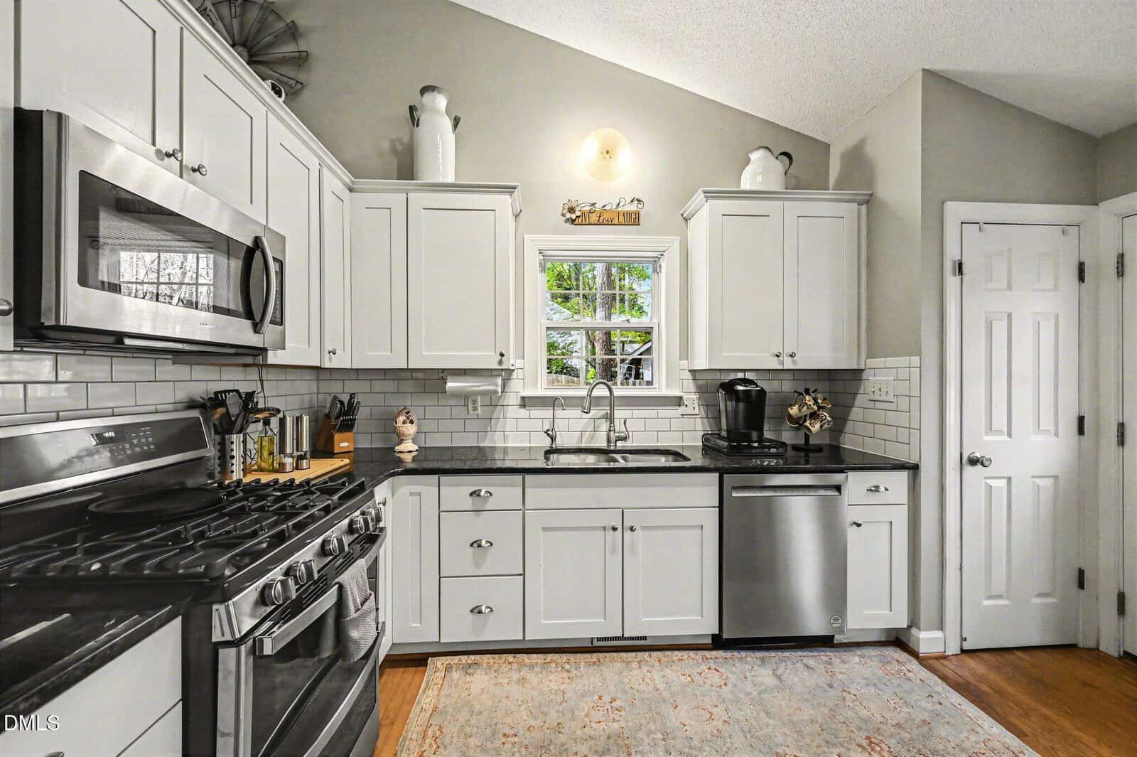 A modern kitchen with white cabinets, stainless steel appliances, a window above the sink, subway tile backsplash, and a patterned rug on a hardwood floor. Decorative items sit on top of the cabinets.