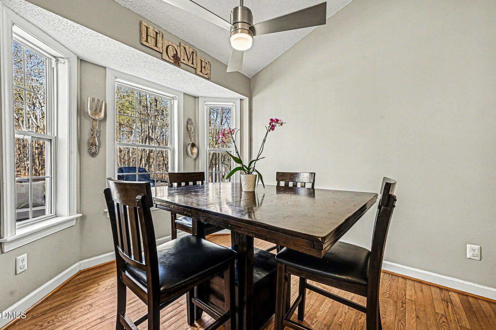 A dining area with a dark wooden table and four chairs, a potted orchid centerpiece, light wood floors, large windows, gray walls, and a ceiling fan with a light. “HOME” sign decorates the wall above the windows.