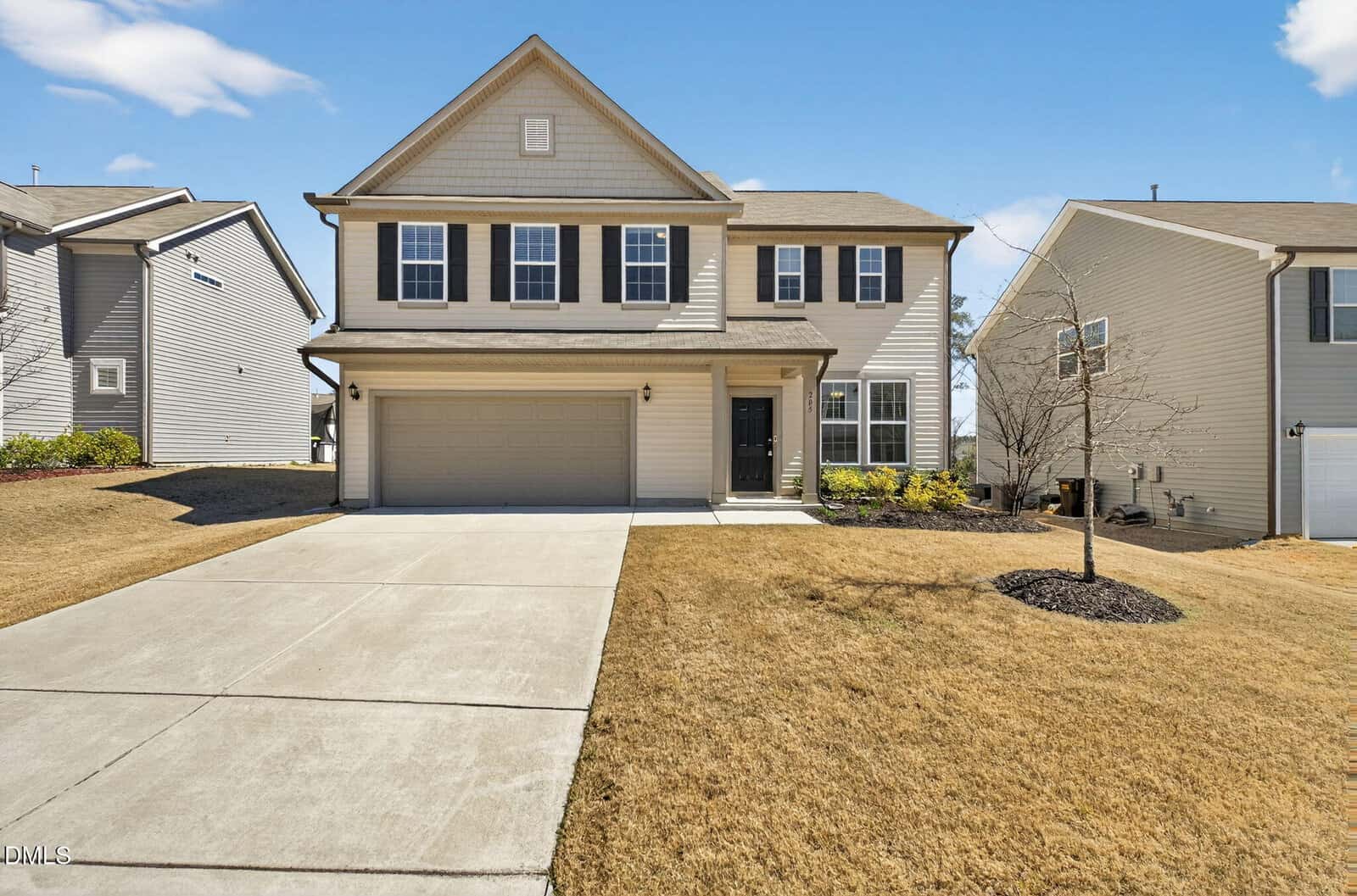 Two-story beige suburban house with black shutters, a double garage, and a concrete driveway. The front yard has dry grass, a small tree, and minimal landscaping. Neighboring houses are visible on both sides.