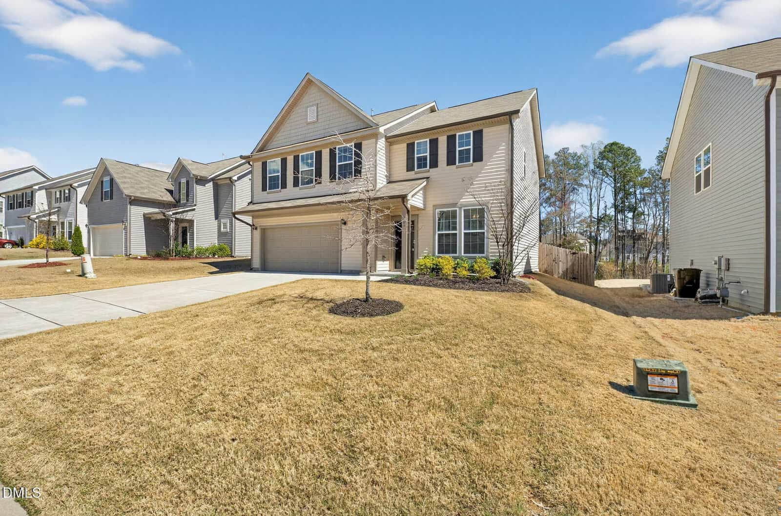 Two-story beige house with brown shutters and a double garage, situated on a sloped lawn in a suburban neighborhood, under a clear blue sky. Neighboring houses and trees are visible in the background.