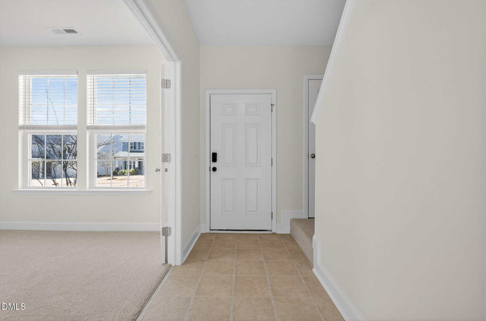 A white front door with a black handle is next to a carpeted staircase. Beige tile flooring and off-white walls are visible, along with a room featuring large windows and natural light.