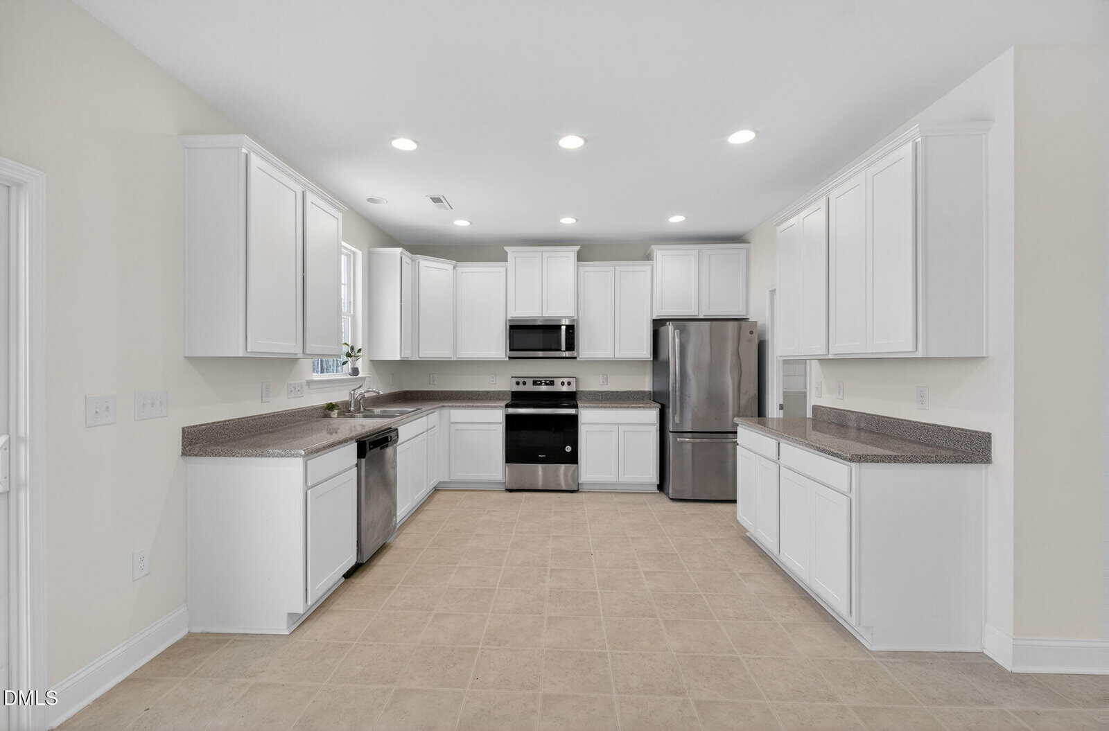 A modern kitchen with white cabinets, stainless steel appliances, beige tile flooring, and gray countertops. The space is well-lit with recessed ceiling lights and a window above the sink.