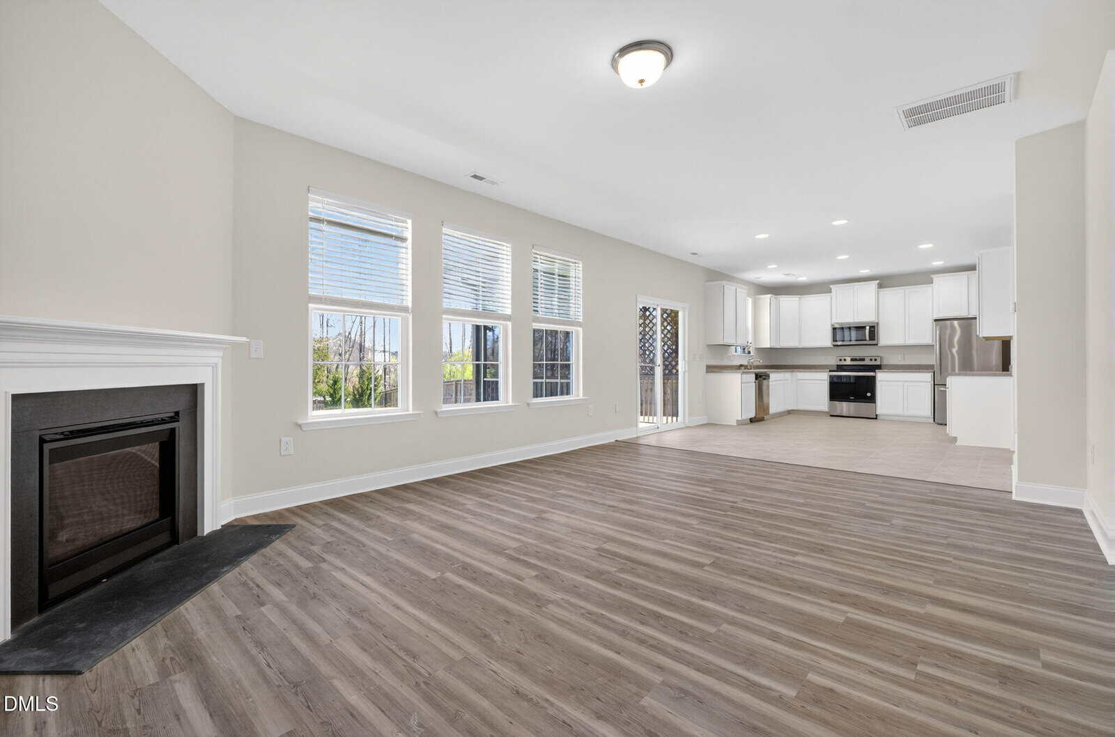 Open-concept living area with light wood flooring, a black-framed fireplace on the left, large windows along the back wall, and a white kitchen with stainless steel appliances in the background.